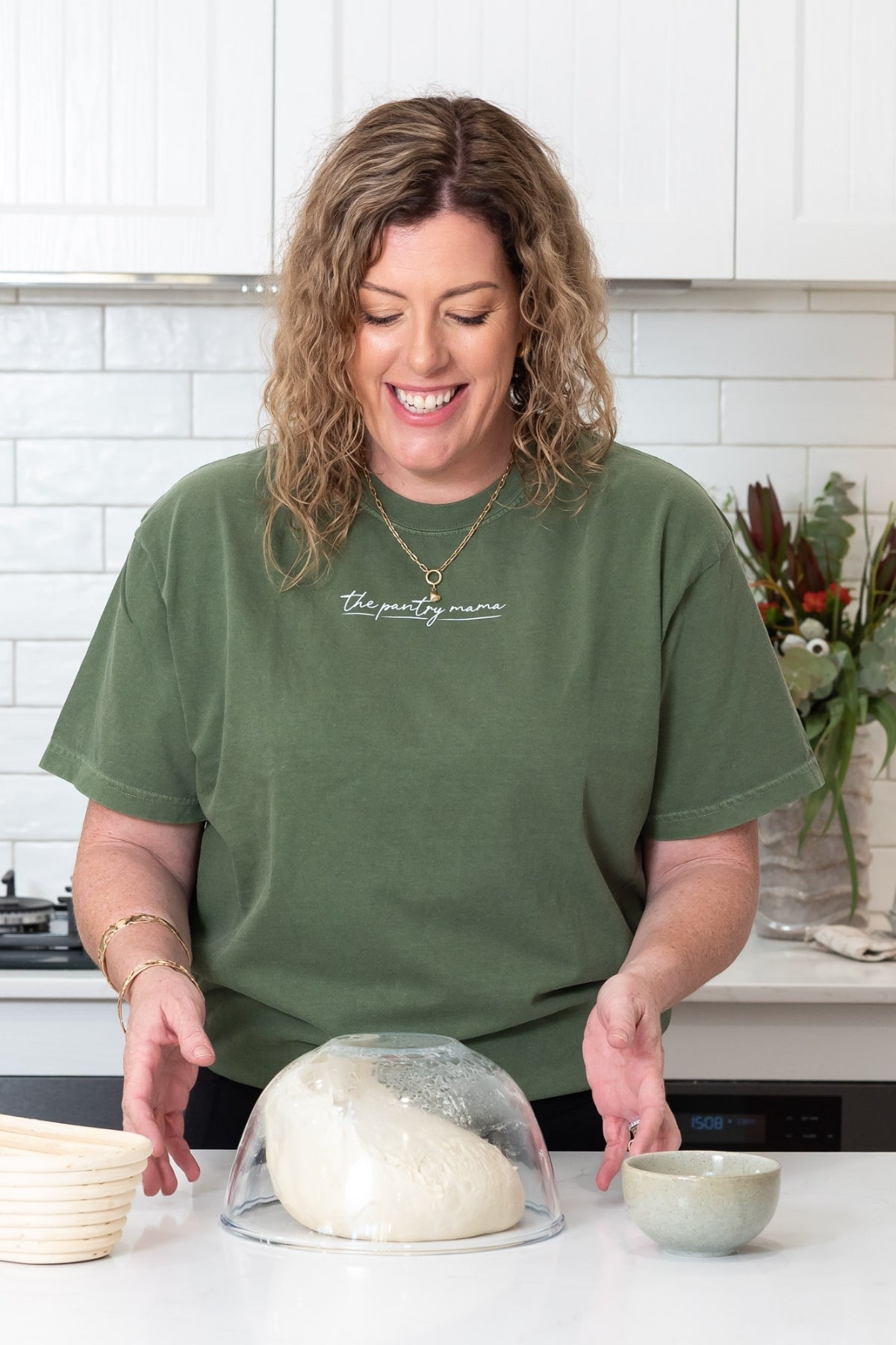 Kate Freebairn, a woman with curly brown hair wearing an olive green t-shirt with 'The Pantry Mama' printed in white script, standing in a bright kitchen behind a glass bowl of sourdough that has been tipped upside down and the dough is falling onto the white counter top.